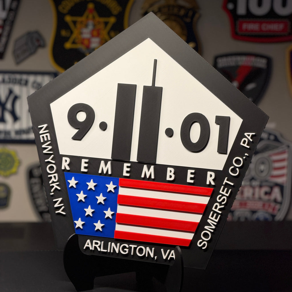 A pentagonal black and white 9/11 memorial plaque with an American flag graphic, displayed on a black stand against a dark background with patches.