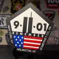 A pentagonal black and white 9/11 memorial plaque with an American flag graphic, displayed on a black stand against a dark background with patches.
