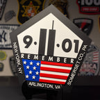 A pentagonal black and white 9/11 memorial plaque with an American flag graphic, displayed on a black stand against a dark background with patches.