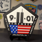A pentagonal black and white 9/11 memorial plaque with an American flag graphic, displayed on a small stand on a dark tabletop. The plaque reads '9-11-01 REMEMBER' and includes location text 'Arlington, VA' and 'New York, NY' and 'Somerset Co.