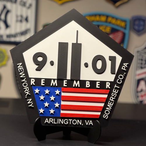 A pentagonal black and white 9/11 memorial plaque with an American flag graphic, displayed on a small stand on a dark tabletop. The plaque reads '9-11-01 REMEMBER' and includes location text 'Arlington, VA' and 'New York, NY' and 'Somerset Co.